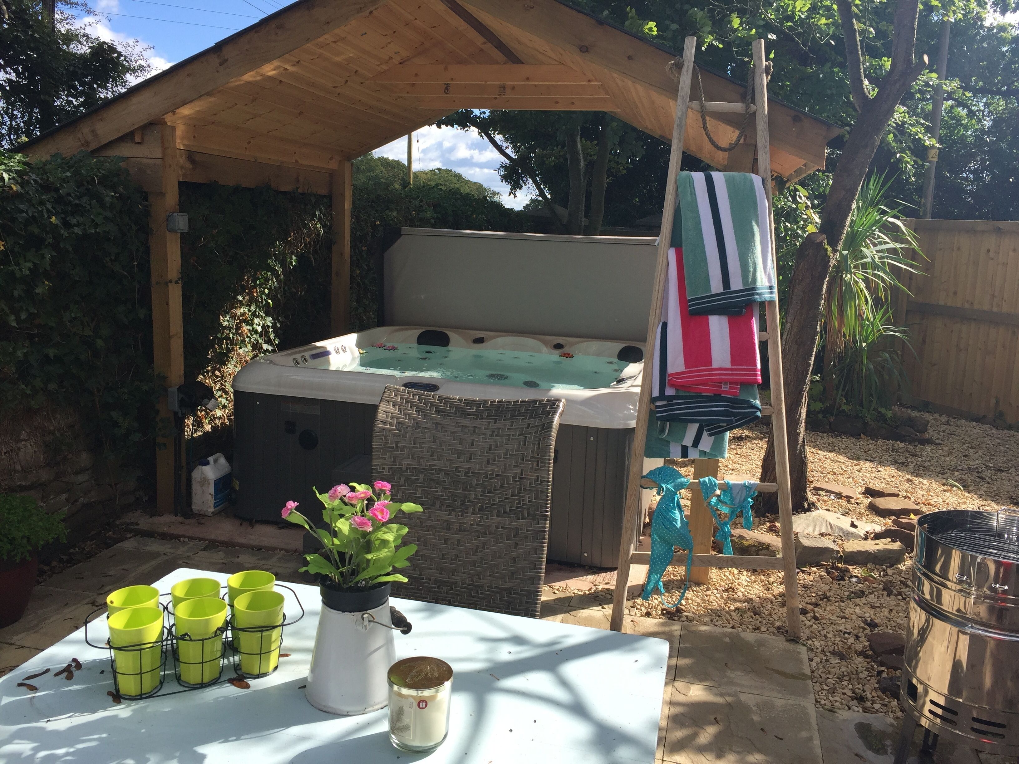 A hot tub under a wooden pergola, surrounded by greenery and a table with drinks and a candle.