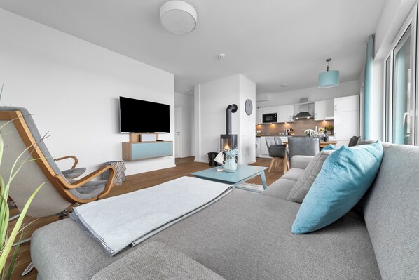 A modern living room with a grey sofa, a wooden chair, a small table, and a TV mounted on the wall. A kitchen is visible in the background. - "Ocean Sunrise" Vacation Apartment with a View of the Baltic Sea – a 3-minute walk from the beach (Olpenitz)
