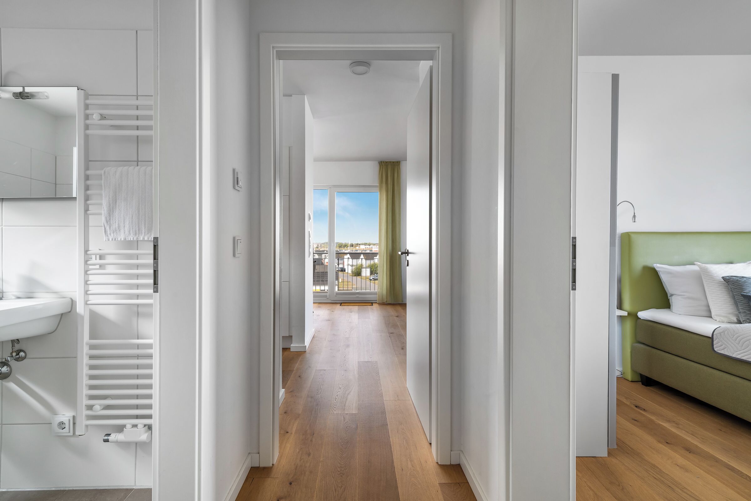 A hotel room with a wooden floor, a green sofa, a white bed, and a view of the city through a balcony.