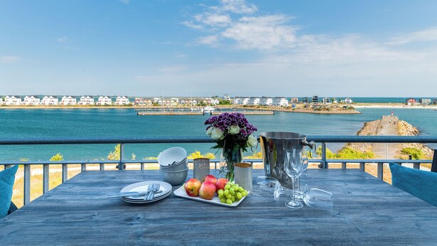 Une table avec une assiette de fruits, un vase de fleurs, un seau et un verre, sur un balcon donnant sur une étendue d’eau et une côte lointaine.
