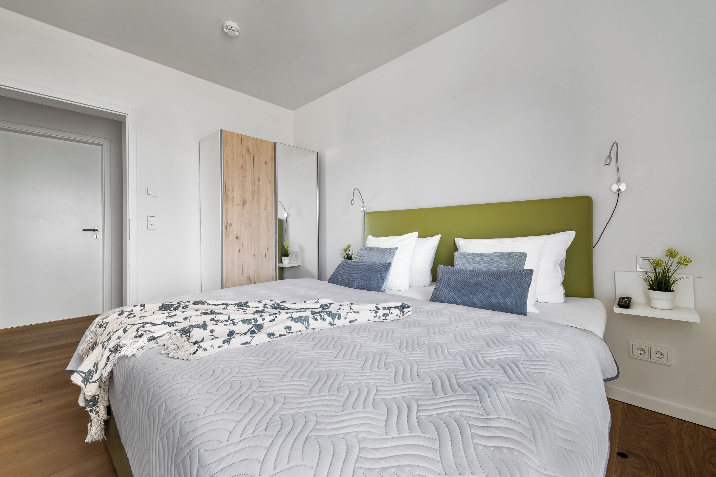 Une chambre moderne avec un grand lit, une tête de lit verte et une table de chevet agrémentée d’une plante.