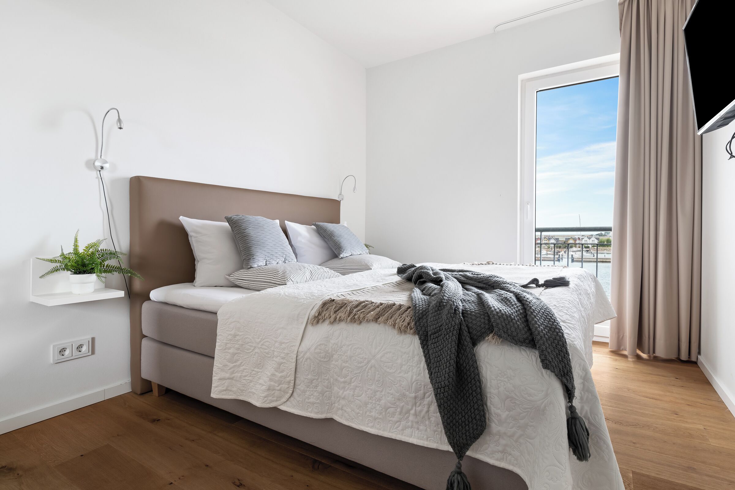 A modern bedroom with a bed, bedside plant, and a view of the outdoors.