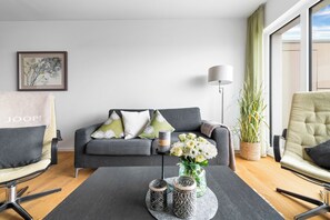 Ein modernes Wohnzimmer mit einem grauen Sofa, zwei Sesseln, einem Couchtisch mit Dekoration, einer Stehlampe und einem großen Fenster mit Vorhängen.