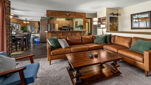 A spacious living room with a large brown leather sectional sofa, a wooden coffee table, and blue armchairs. A kitchen area is visible in the background with a bar counter and seating. - Close to the Slopes! Pool & Hot Tubs! Winter Shuttle! (Steamboat Springs)