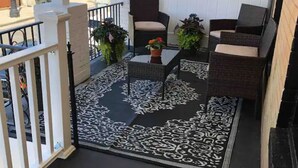 Un balcón con mobiliario de mimbre, alfombra estampada y plantas en macetas.