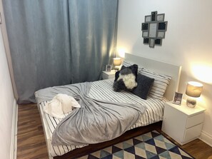 Una cama bien tendida con ropa de cama a rayas, una manta gris y una almohada blanco y negro. Hay dos mesitas de noche con lámparas, un reloj de pared y un cuadro enmarcado. Una cortina azul cubre una ventana, y una alfombra estampada está en el suelo.
