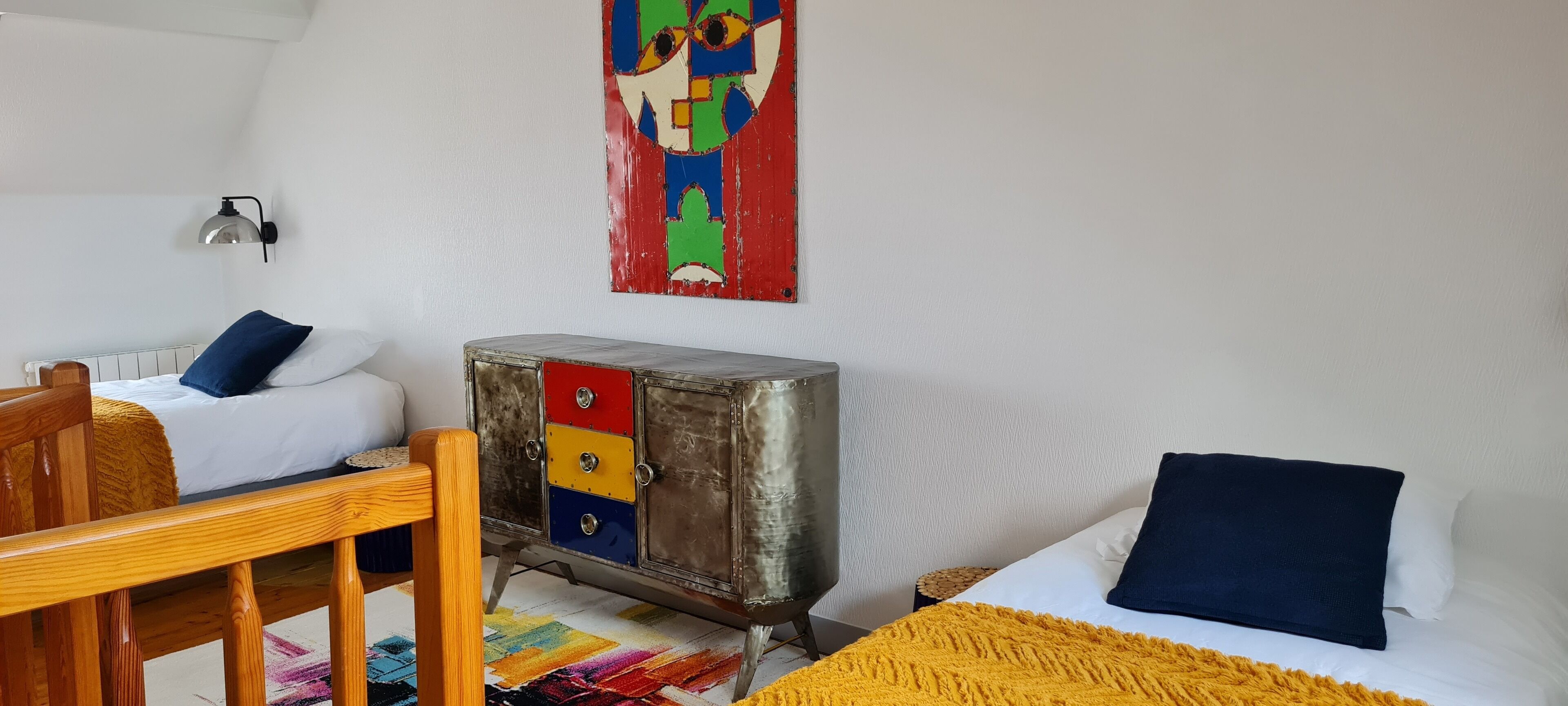 Ein Schlafzimmer mit zwei Betten, einem Holzhocker, einem Metallschränkchen mit Schubladen, einem farbenfrohen Teppich und einem Wandbild.