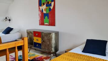 Ein Schlafzimmer mit zwei Betten, einem Holzhocker, einem Metallschränkchen mit Schubladen, einem farbenfrohen Teppich und einem Wandbild.