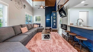 A modern living room with a grey sectional sofa, a pink shag rug, and a small wooden table. There is a kitchen area with a blue accent wall, a bar counter, and stools.