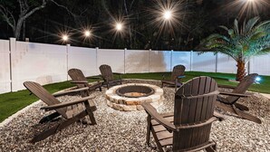 An outdoor seating area with Adirondack chairs arranged around a circular fire pit, surrounded by gravel and a white fence.