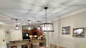A spacious dining area with a long wooden table and matching chairs, a kitchen in the background, and pendant lighting.