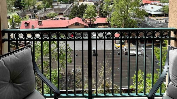 A balcony with a view of a town surrounded by hills, featuring a red-roofed building and a parking lot.