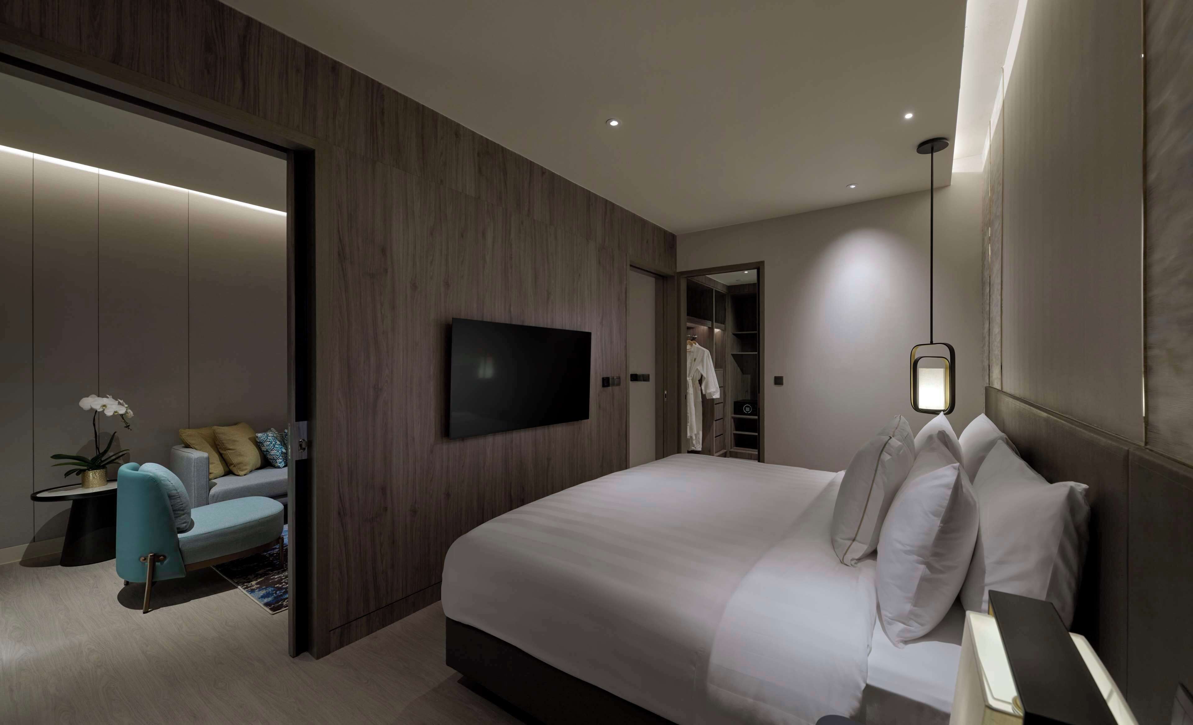 Una habitación de hotel moderna con una cama grande, televisión montada en la pared, armario y zona de estar con una silla y una mesita.