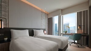 Een hotelkamer met twee bedden, een bureau, een stoel, een raam met uitzicht op de stad en een lamp.