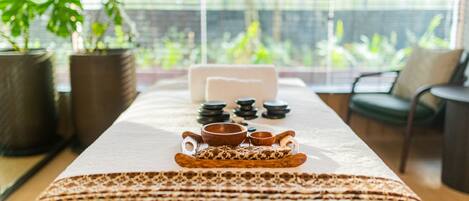 A long dining table with a tray of food and bowls, potted plants, and a view of greenery through large windows.