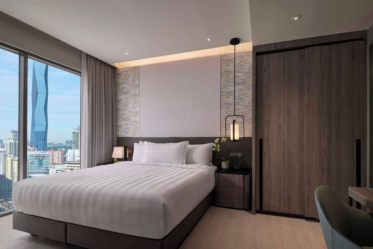 Une chambre d’hôtel moderne dotée d’un grand lit, d’une table de chevet avec une lampe, d’un petit bureau avec une chaise et offrant une vue sur le panorama urbain.