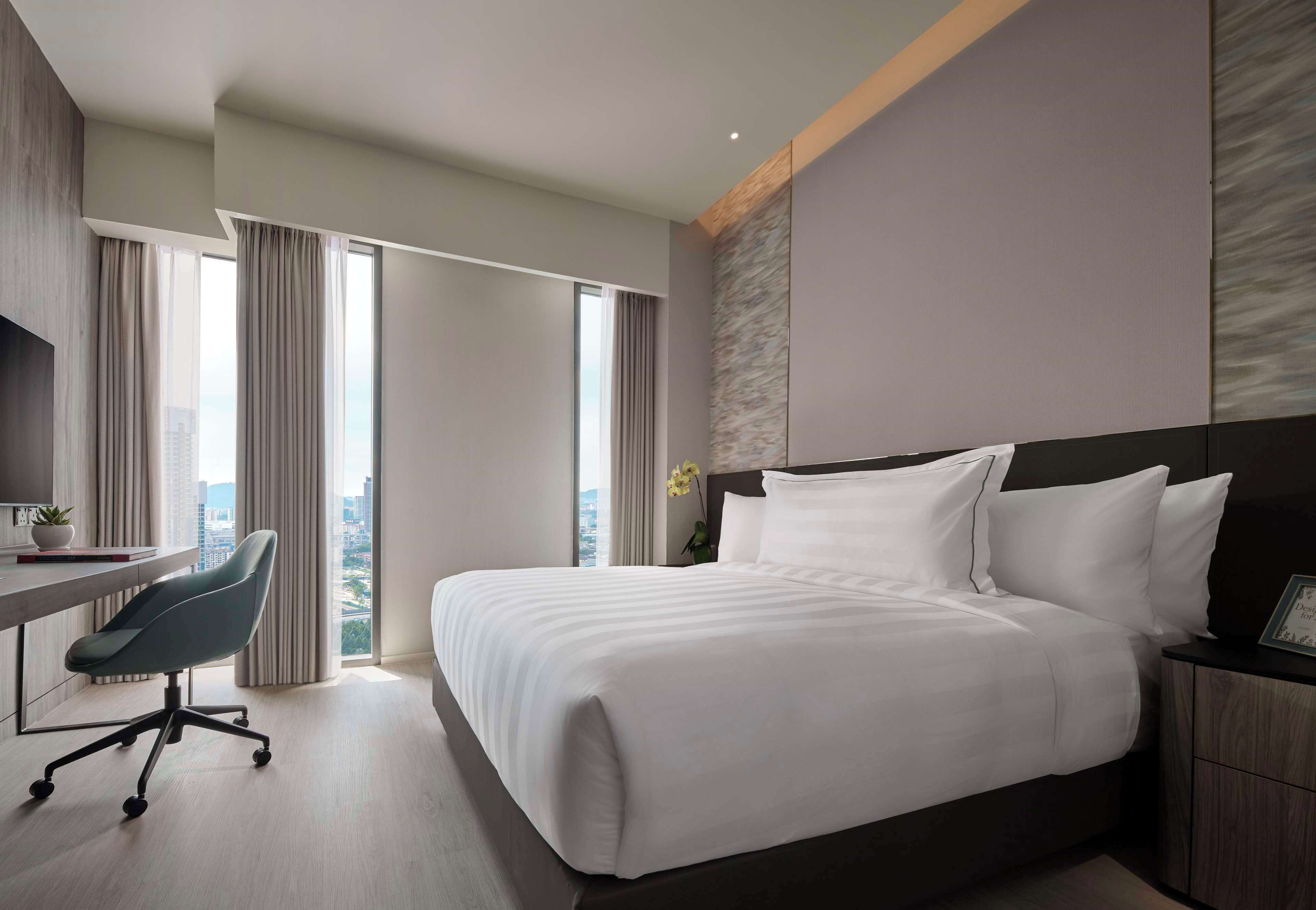 A modern hotel room with a large bed, a desk with a chair, and a view of the city through large windows.