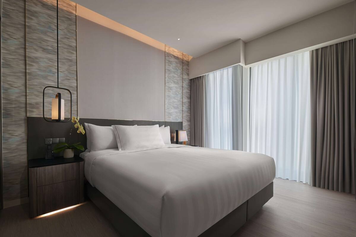 Une chambre d’hôtel moderne avec un grand lit, une table de chevet avec une lampe, une plante en pot et une grande fenêtre avec des rideaux.