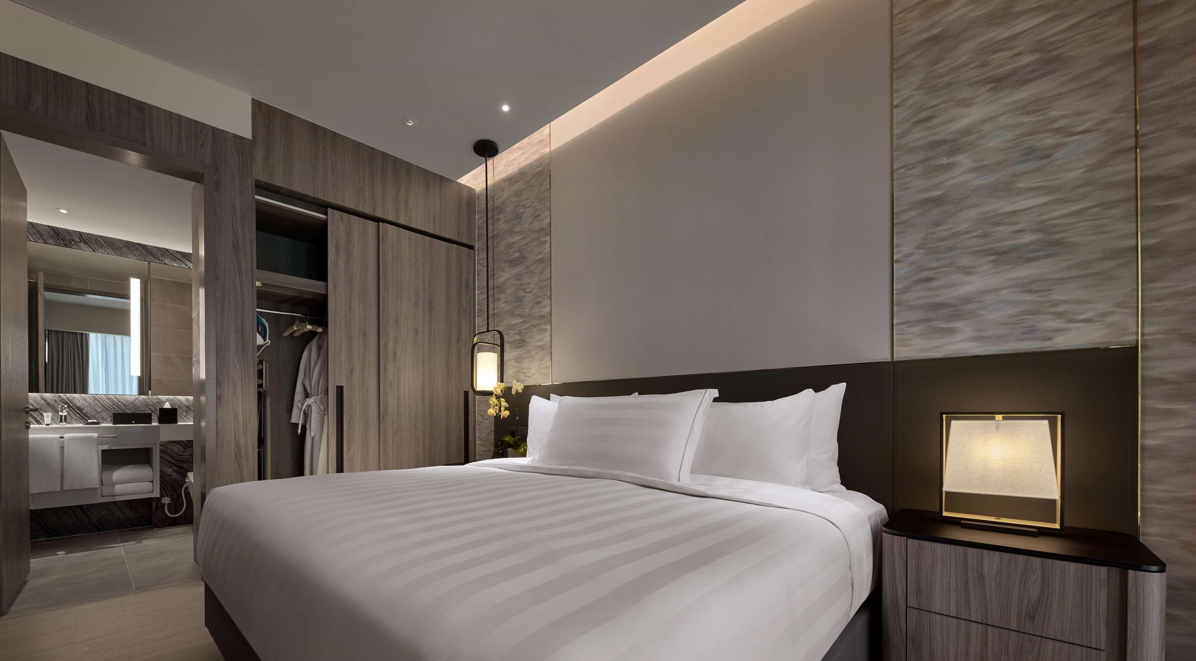 Una habitación de hotel moderna con una cama grande, un vestidor, un baño con tocador y una mesita de noche con lámpara.