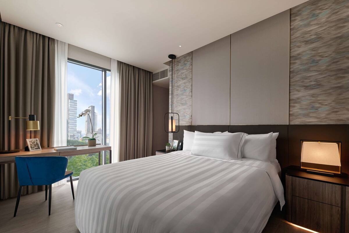 Une chambre d’hôtel moderne dotée d’un grand lit, d’un bureau avec une chaise, d’une table de chevet et offrant une vue sur les toits de la ville grâce à de grandes fenêtres.