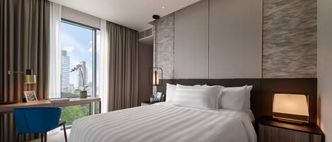A modern hotel room with a large bed, a desk with a chair, a nightstand, and a view of the city skyline through large windows.