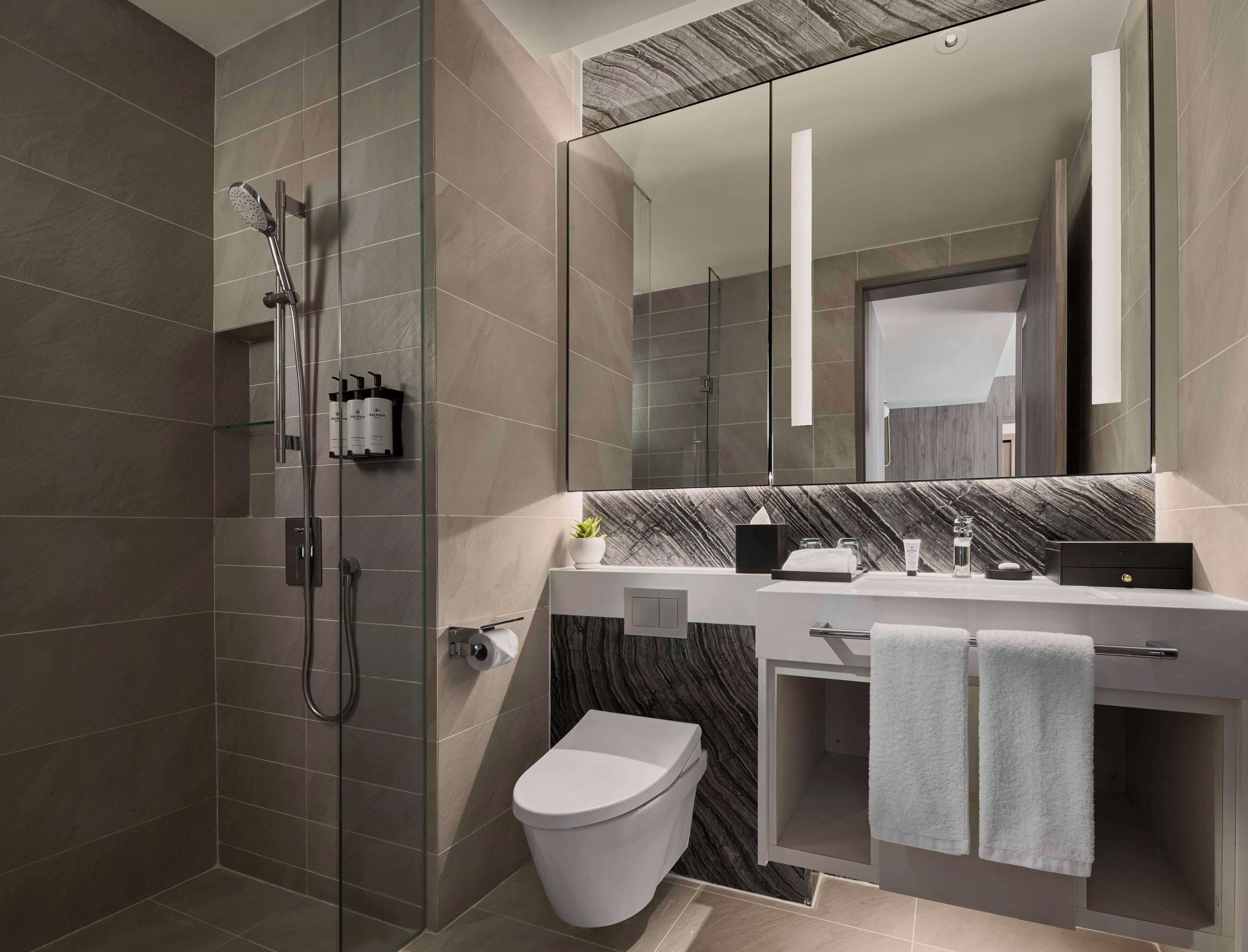 Un baño moderno con ducha de vidrio, inodoro blanco, salpicadero de mármol y un espejo grande.