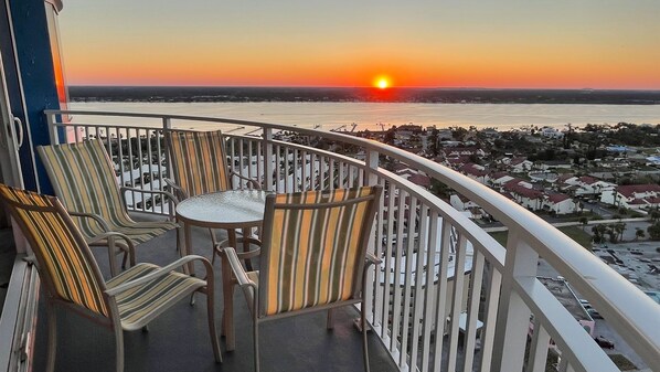 A balcony with chairs and a table, overlooking a sunset over a body of water and a residential area.