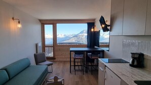Ett kompakt bostadsutrymme med en soffa, ett matbord med stolar, ett köksområde med en kaffebryggare och ett stort fönster med utsikt över bergen.