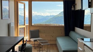 Ett kompakt rum med trägolv, ett matbord med stolar, en soffa, ett litet bord och en tv. Ett stort fönster ger utsikt över bergen.