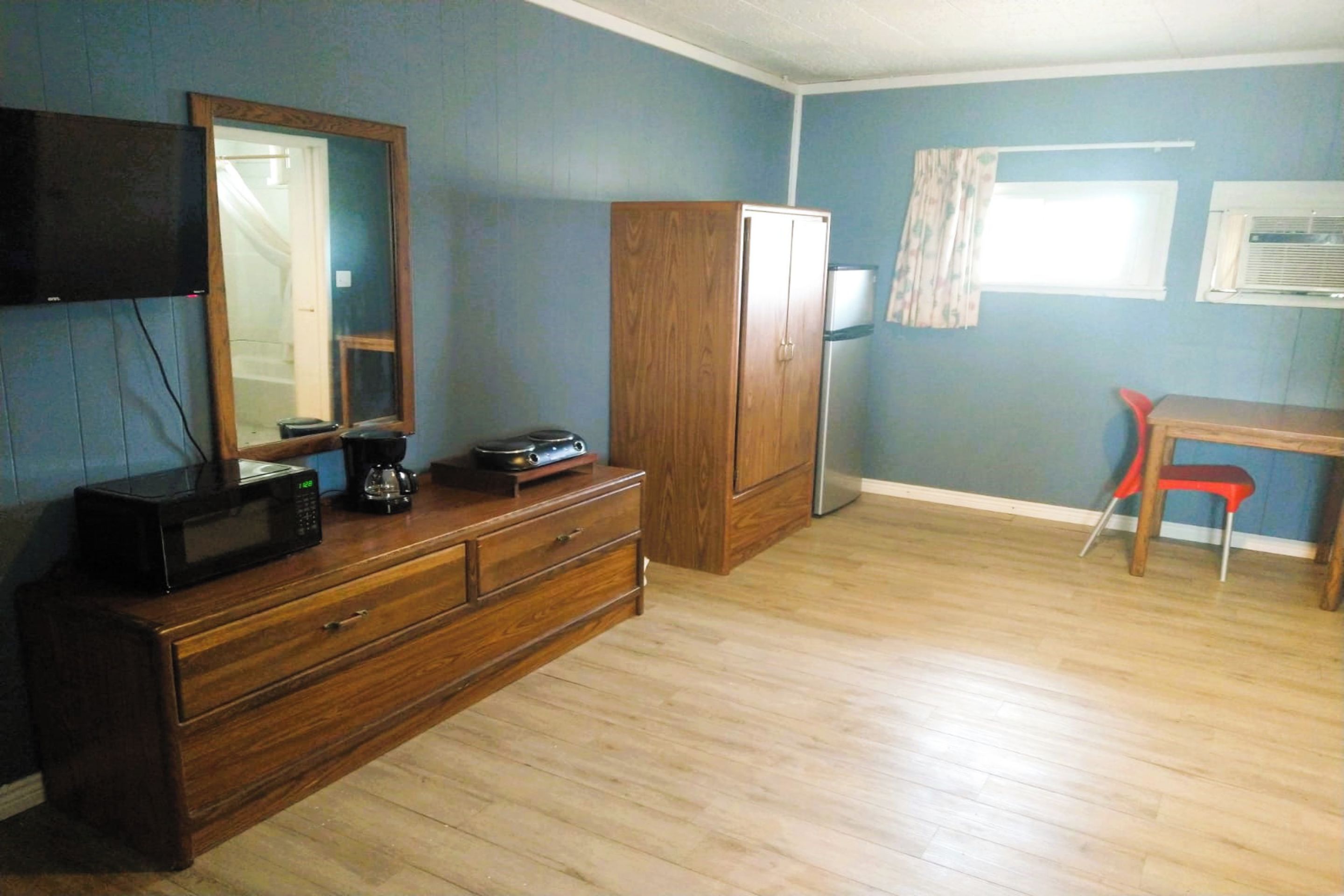 A room with a wooden dresser, a flat-screen TV mounted on the wall, a wooden cabinet, a red chair, and a window with a curtain.