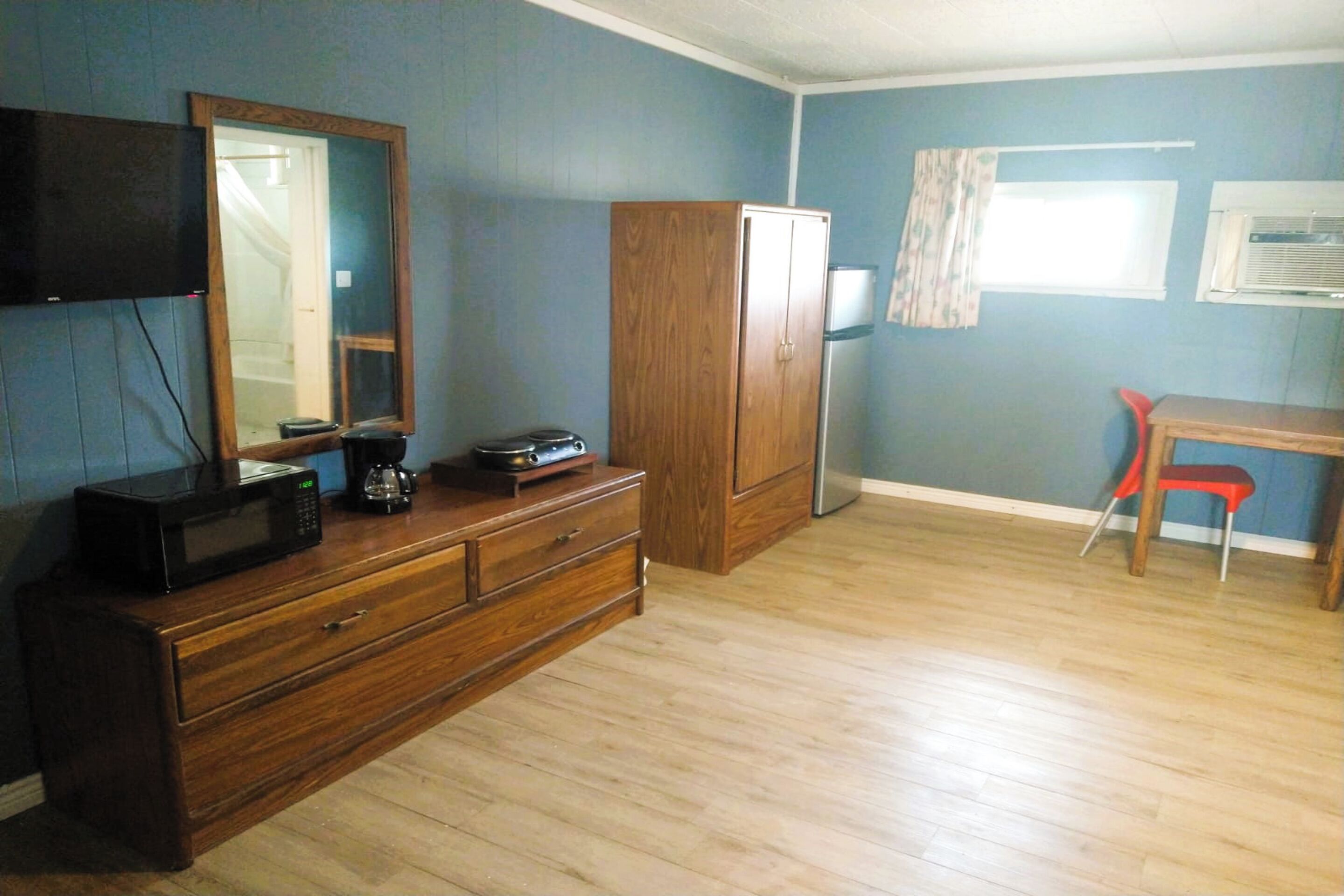 A room with a wooden dresser, a flat-screen TV mounted on the wall, a wooden cabinet, a red chair, and a window with a curtain.