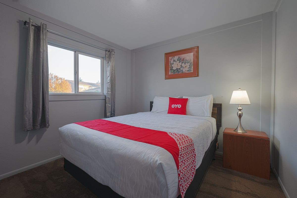 A hotel room with a large bed, a nightstand with a lamp, a window with curtains, and a painting on the wall.