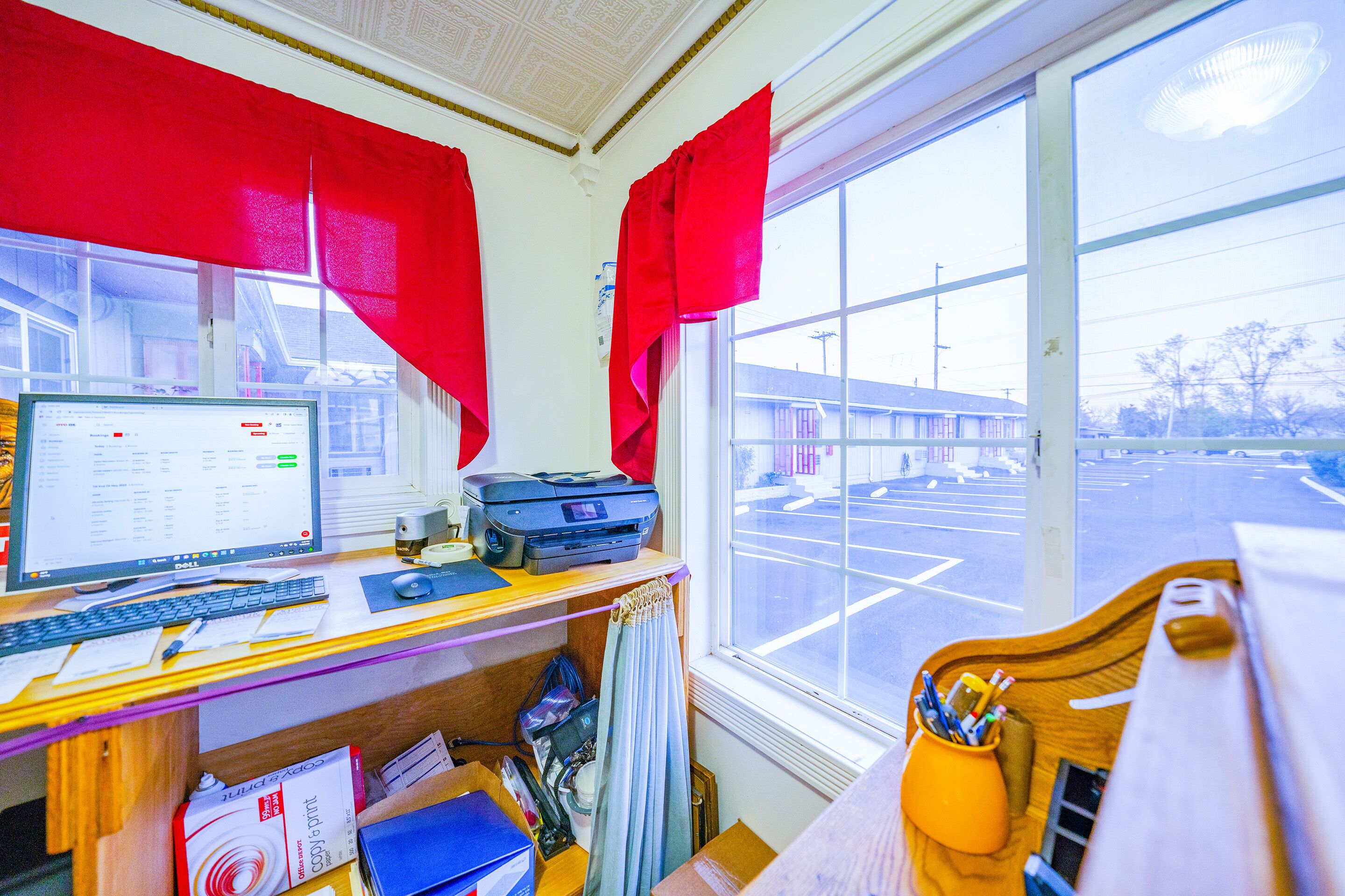 A room with a desk, computer, printer, and a window with a view of a parking lot.