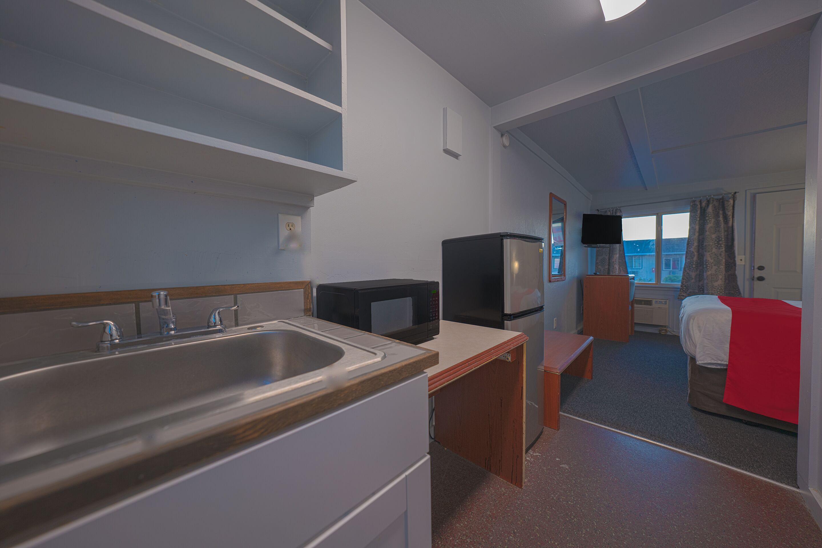 A compact hotel room with a kitchenette, a bed with a red blanket, a television, and a window with curtains.