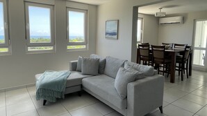 A living room with a grey sofa, a dining table with chairs, and large windows with a view. - Ocean view apartment- close to beaches and Yunque (Luquillo)