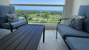 A balcony with two blue cushioned chairs and a wooden table, overlooking a green landscape and the ocean. - Ocean view apartment- close to beaches and Yunque (Luquillo)