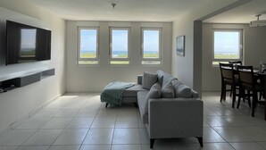 A modern living room with a grey sofa, a flat-screen TV mounted on the wall, and three windows offering a view of the outdoors. - Ocean view apartment- close to beaches and Yunque (Luquillo)