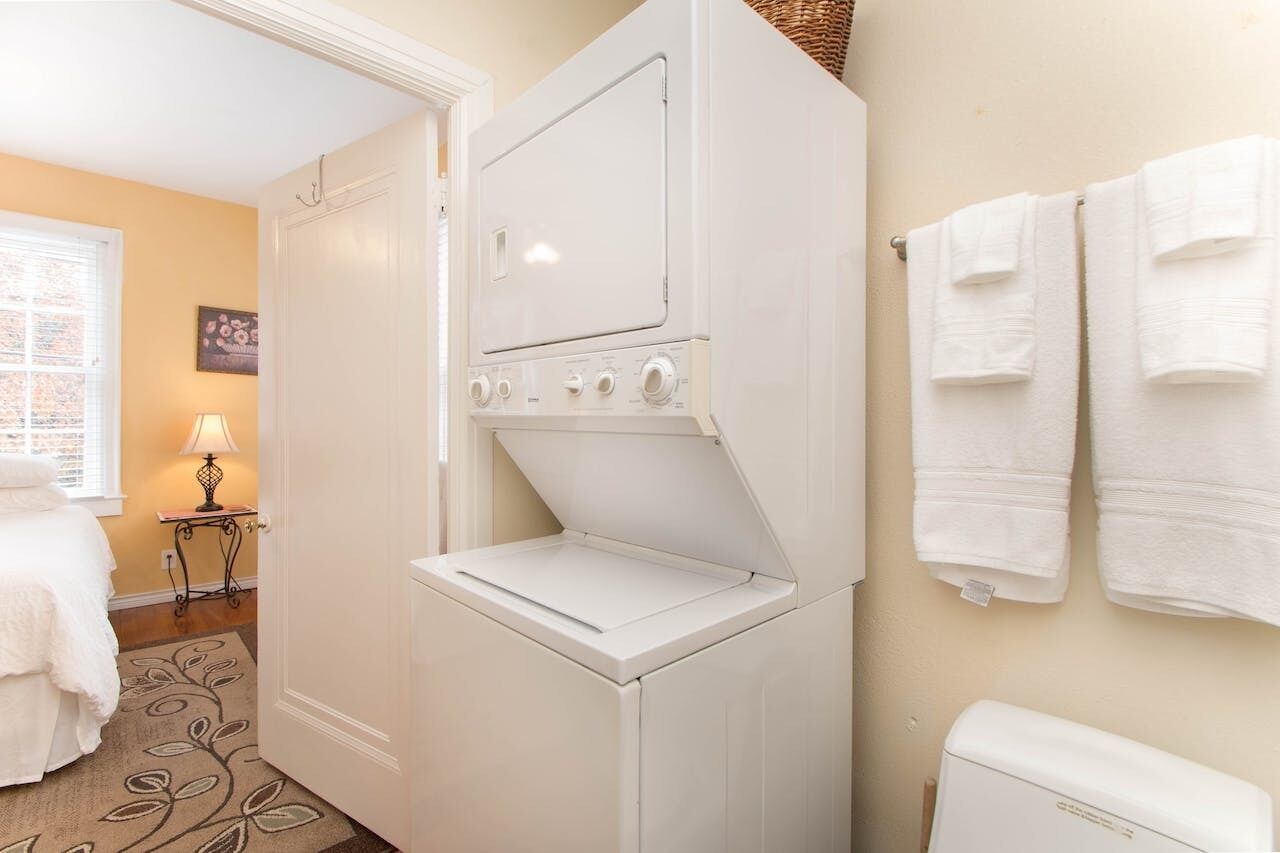 Une petite buanderie équipée d’un lave-linge et d’un sèche-linge à chargement frontal, un lit avec une couette blanche, une petite table avec une lampe et une fenêtre avec des stores.