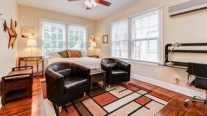 A bedroom with a bed, two chairs, a desk, a ceiling fan, and large windows with blinds.