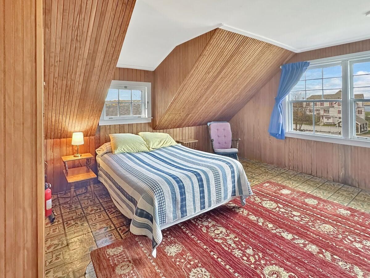 A bedroom with a sloped ceiling, a bed with a striped bedspread, a red patterned rug, a nightstand with a lamp, and a window with curtains.