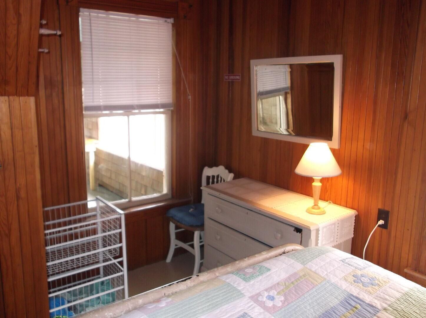 Een kleine, compacte slaapkamer met houten wanden, een enkel bed, een dressoir, een stoel, een raam met zonwering en een spiegel.