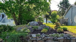 Um jardim paisagístico com muro de pedra, arbustos diversos e cerca de piquetes brancos.