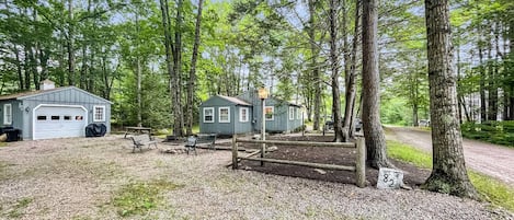 A rustic cabin with a garage, a small shed, and a picnic table in a wooded area.