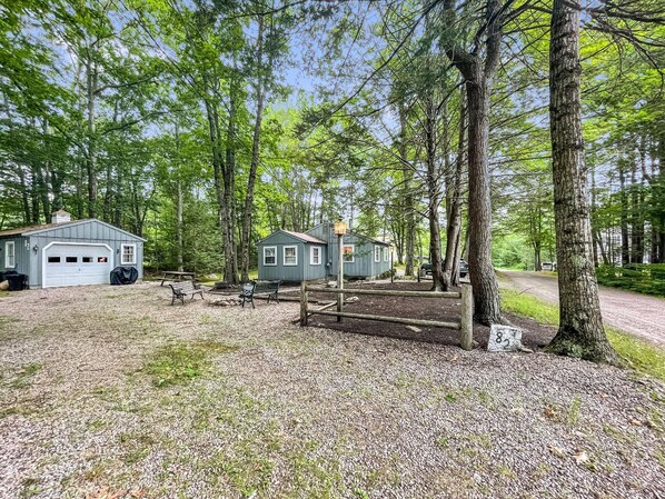 A rustic cabin with a garage, a small shed, and a picnic table in a wooded area.