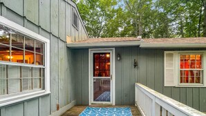 A covered porch with a blue mat featuring white anchors, a wooden deck, and a door leading to an interior room with a visible window and light fixture. - Dewey Cottage (York Beach)