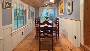 A dining area with a wooden table and chairs, a ceiling fan, and large windows with a view of the outdoors. - Dewey Cottage (York Beach)