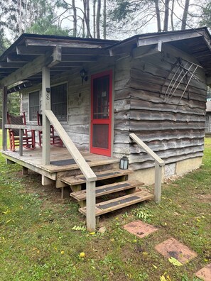 A rustic cabin with a porch, a red door, and a set of wooden stairs leading to the entrance.