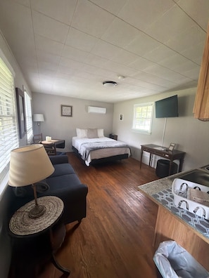 A compact hotel room with a bed, a desk, a chair, a small table, a lamp, and a television.