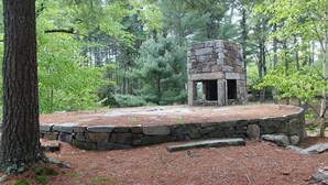 A stone-built outdoor fireplace surrounded by a stone wall, set in a wooded area with pine trees. - Lighthouse - Q991 (Cape Neddick)