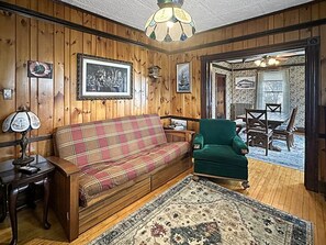 Ein Zimmer mit Holzwänden, einem Karo-Sofa, einem grünen Sessel, einem gemusterten Teppich und gerahmten Bildern an den Wänden.